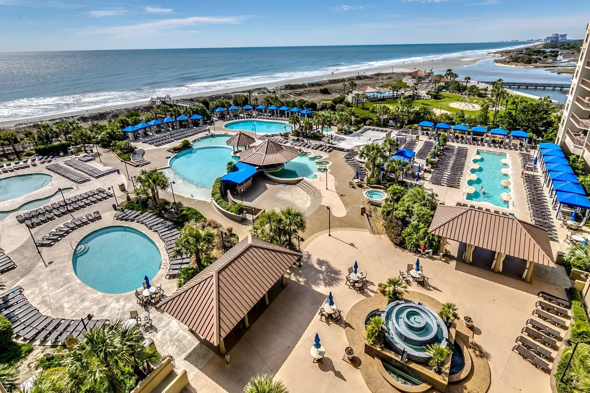 A vibrant, resort-style pool complex featuring multiple swimming pools, palm trees, poolside cabanas, seating areas, and a beachfront view. The image captures both lounge areas and inviting water spaces, evoking a sense of luxury and fun.
