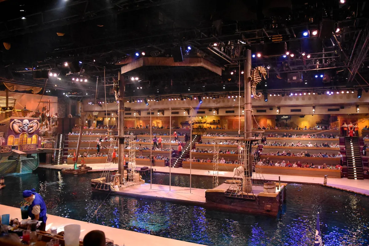 This energetic image displays a live performance with a grand pirate ship set on a water stage, surrounded by an enthusiastic audience. The vibrant lighting and themed decor hint at the unique entertainment offered nearby.