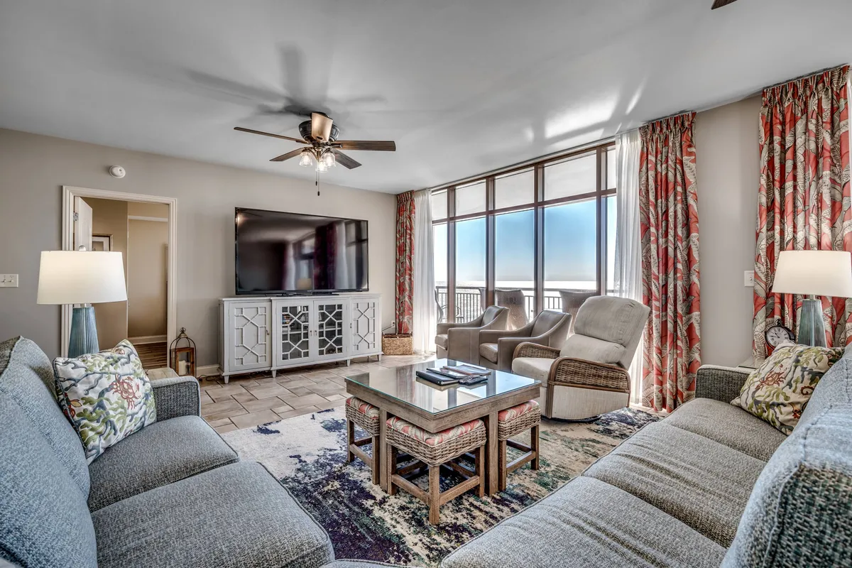 The image showcases a spacious, inviting living area with plush seating arranged around a stylish coffee table. Floor-to-ceiling windows fill the room with natural light and offer expansive views—perfect for sunset watching. Elegant decorative curtains frame the windows, creating a cozy yet upscale atmosphere ideal for gathering with family or friends.