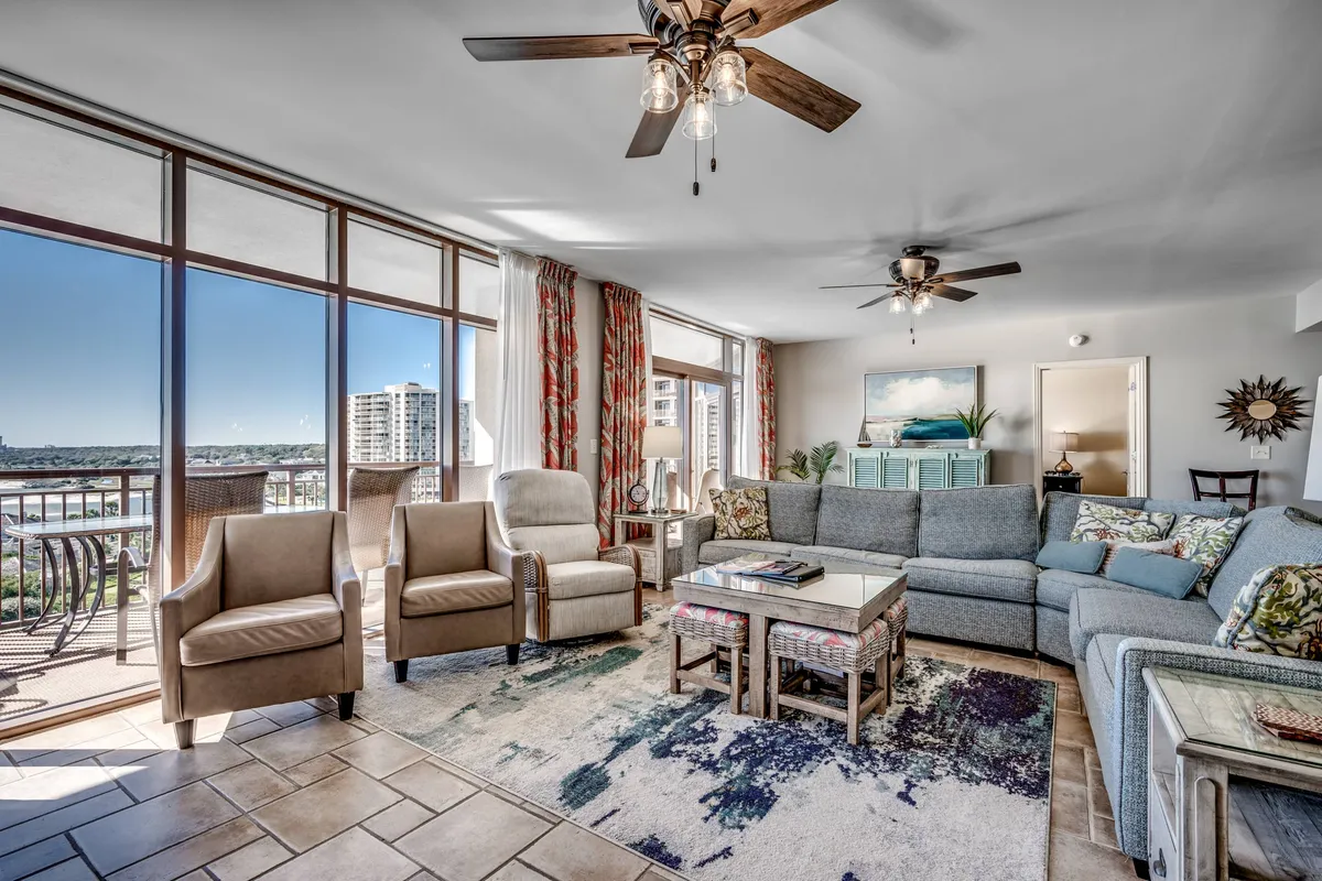 A spacious living area with large windows overlooking an outdoor scene, colorful sofa, stylish coffee table, and artwork on the wall. The room is filled with natural light, offering views that hint at the beach or pool just beyond.