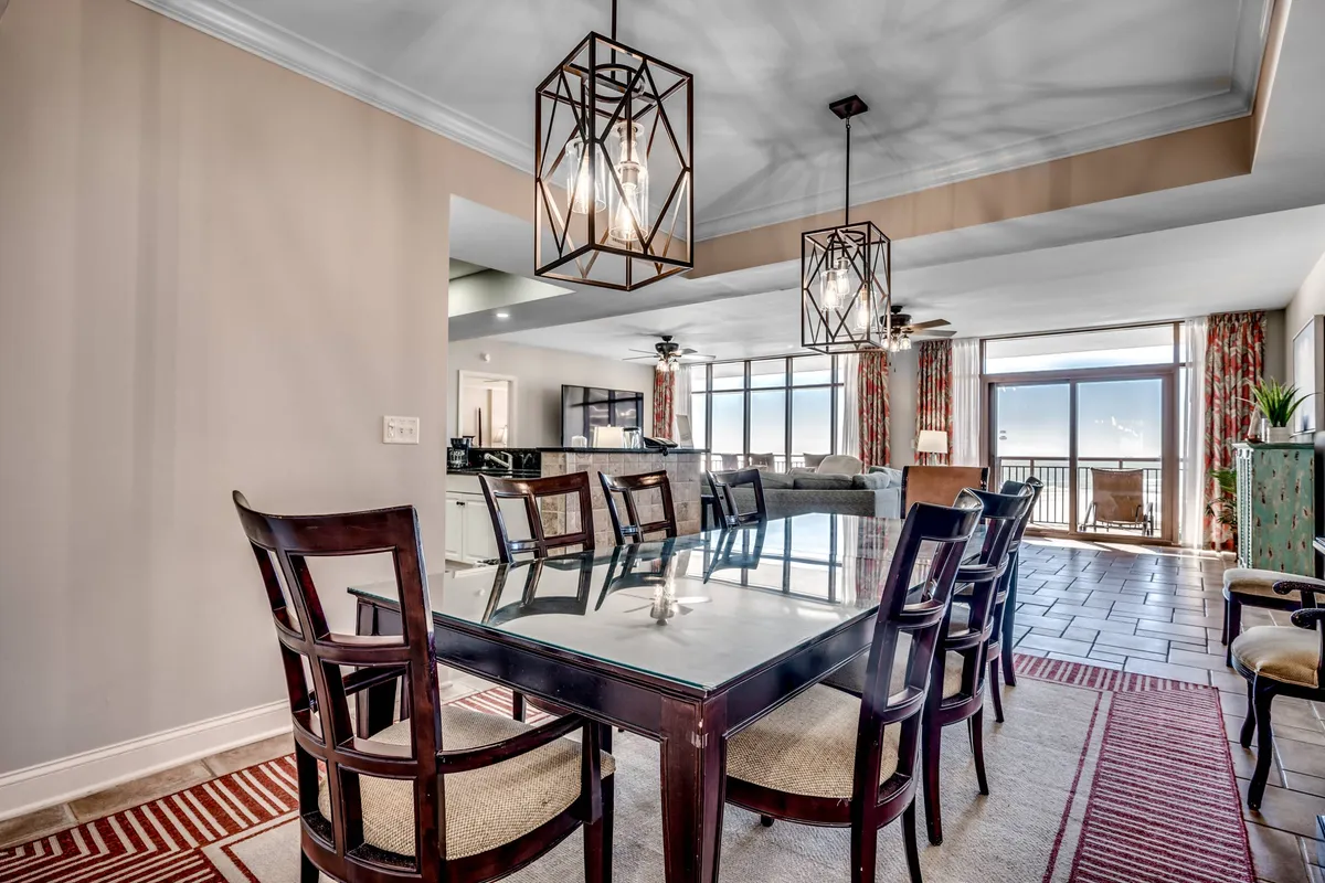 A modern dining area with a large table, elegant hanging light fixtures, and expansive windows that let in abundant natural light. A comfortable couch sits in the background, highlighting the seamless open-concept flow between dining and living spaces.
