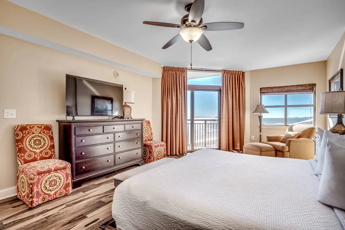 A bright, comfortable bedroom featuring a neatly made bed, a large window with a scenic view, a dresser with a television, and an armchair for relaxation. The room feels airy and well-organized, highlighting both comfort and ample storage.