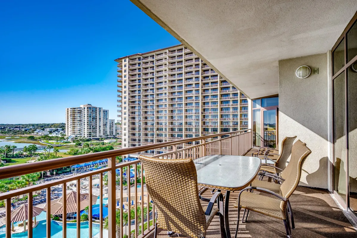 This exterior image captures a private balcony with comfortable seating overlooking the resort’s pool area and surrounding buildings. The view suggests an inviting transition between indoor luxury and outdoor leisure, with the scene bathed in soft natural light, ideal for relaxing with a drink or enjoying the sunset.