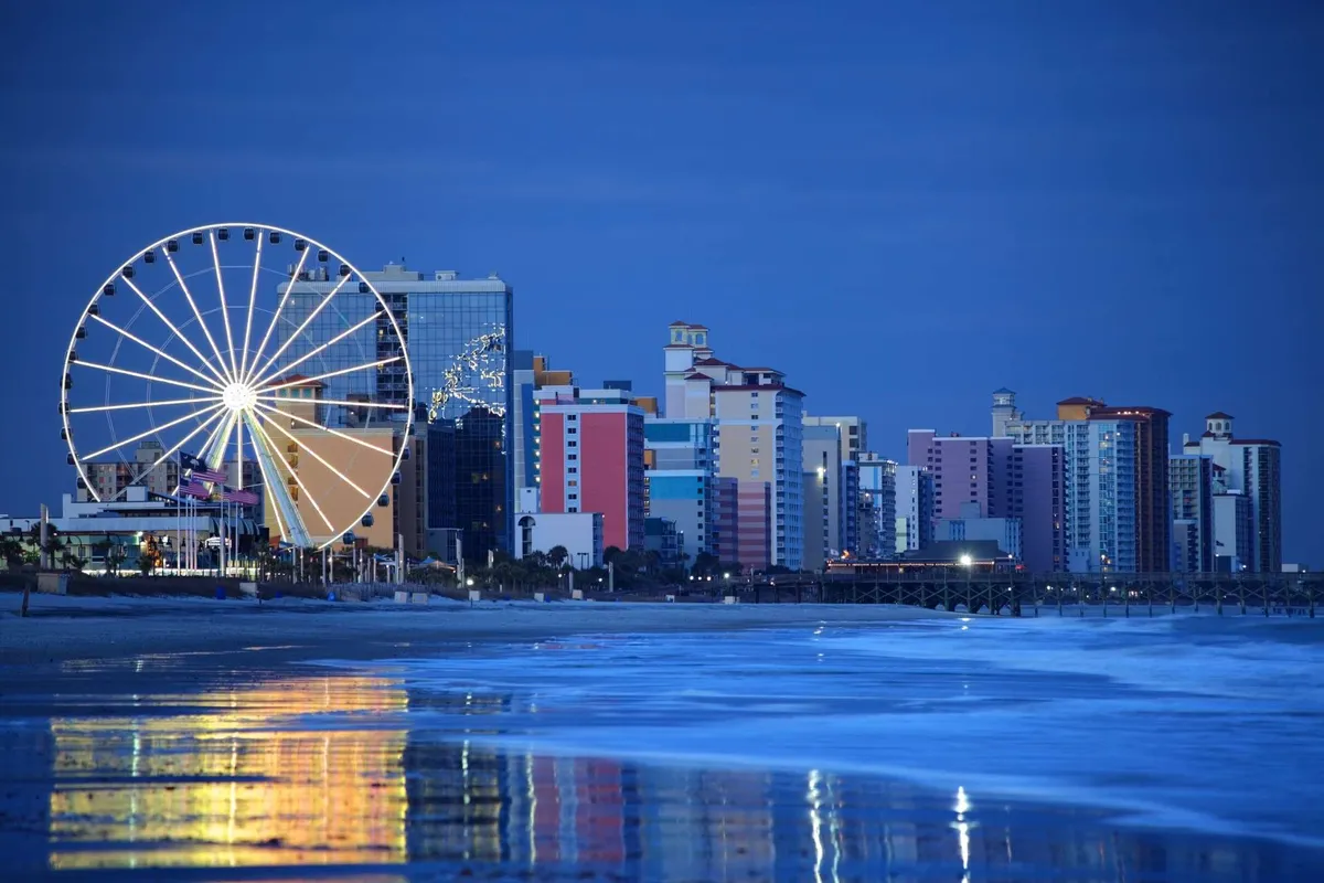 A vibrant exterior shot showcasing the iconic ferris wheel, colorful buildings, and reflections on the water, evoking the lively nightlife and entertainment of the Grand Strand. The setting transitions from day to night, suggesting both playful beachside activities and evening adventures.