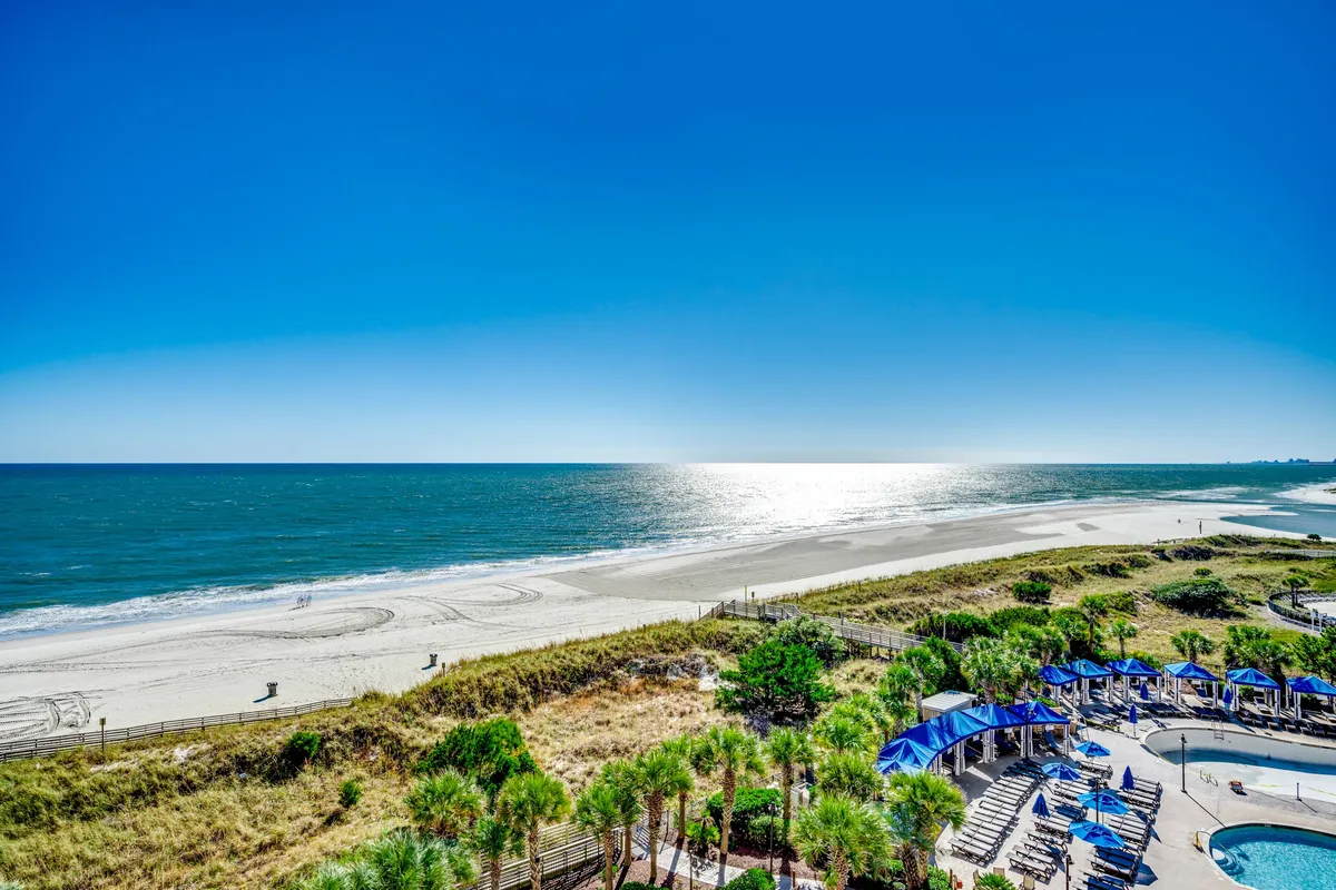 A sun-drenched beach scene featuring gentle ocean waves, golden sands, clusters of blue umbrellas, and lounge chairs. Palm trees add to the inviting, tropical atmosphere, perfect for both relaxation and active beach days.