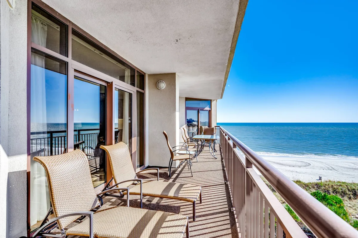This image captures a serene balcony view overlooking the expansive beach and ocean. Comfortable seating on the balcony invites guests to relax while soaking in the natural beauty, with the pristine sand and gentle waves visible just beyond.