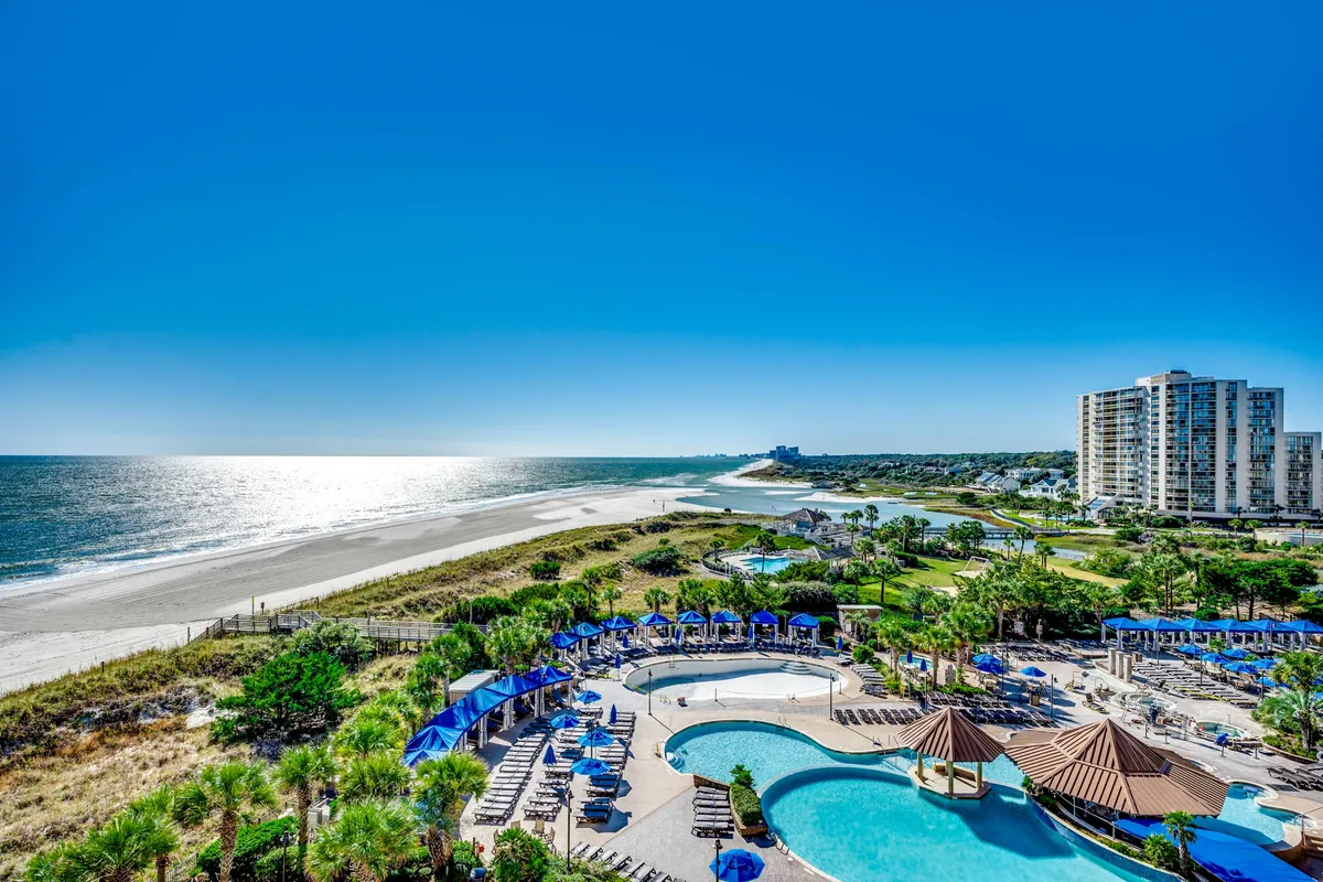 A wide view encompassing the sparkling swimming pool, nearby beach, and the ocean stretching into the distance. Lounge chairs dot the poolside, with buildings and palm trees in the background, evoking a sense of resort-style relaxation and outdoor enjoyment.