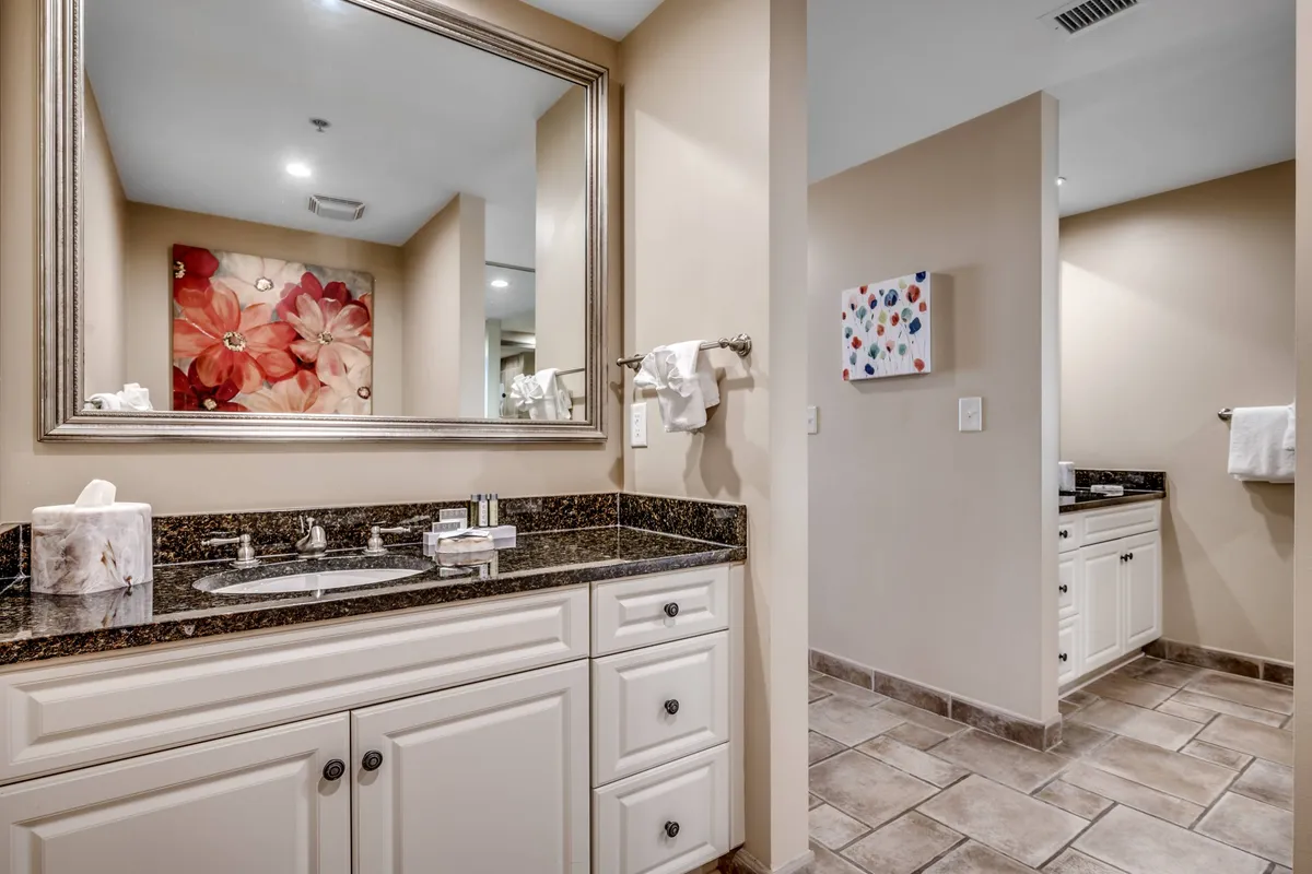 A calm, elegant spa-like bathroom with a large mirror, flower artwork, granite countertop, and sink. The soft lighting and decor suggest a focus on relaxation, grooming, and self-care routines.