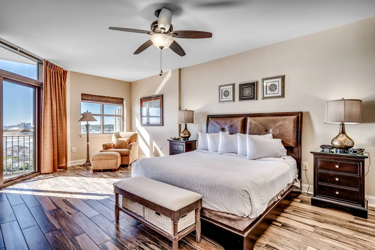 A serene bedroom with a king-sized bed dressed in plush, inviting linens. Natural light pours through a large window that offers a peaceful view, while a comfortable armchair and tasteful decorative artwork create a tranquil, personalized retreat. The room appears spacious and well-appointed, ideal for unwinding after a busy day.